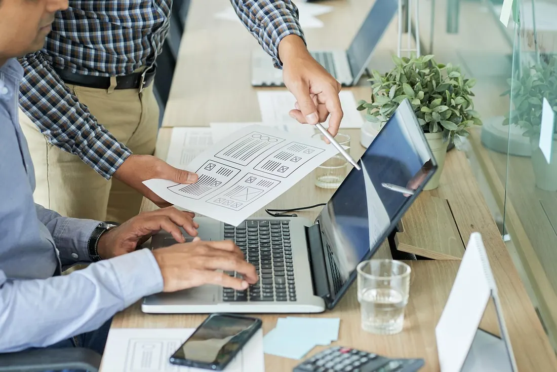 Two people at a desk reviewing printed website wireframes next to a laptop